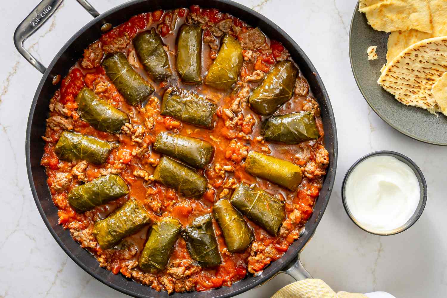A skillet with dolmas cooked in tomato sauce and ground beef, served with pita bread and yogurt
