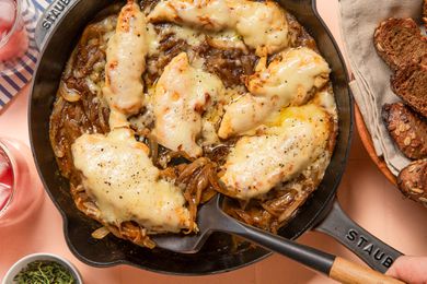 french onion chicken in a skillet at a table setting with glasses, a bowl of bread, and a small bowl of herbs