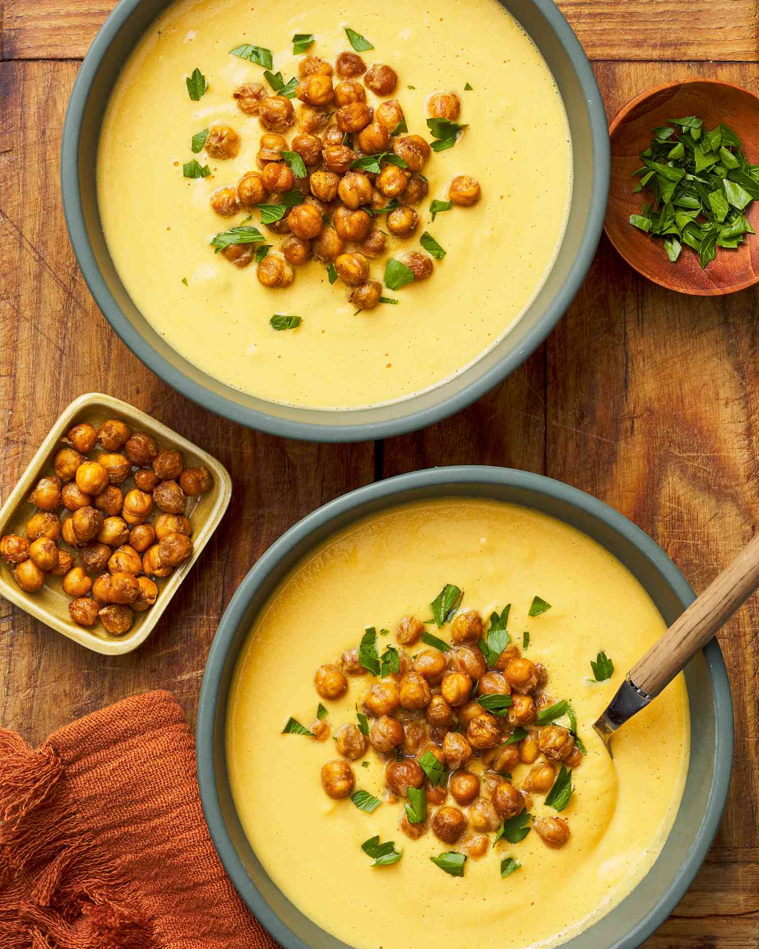 Bowls of hummus next to a small bowl of roasted chickpeas, another small bowl with chopped parsley, and a burnt orange kitchen towel, all on a wooden table