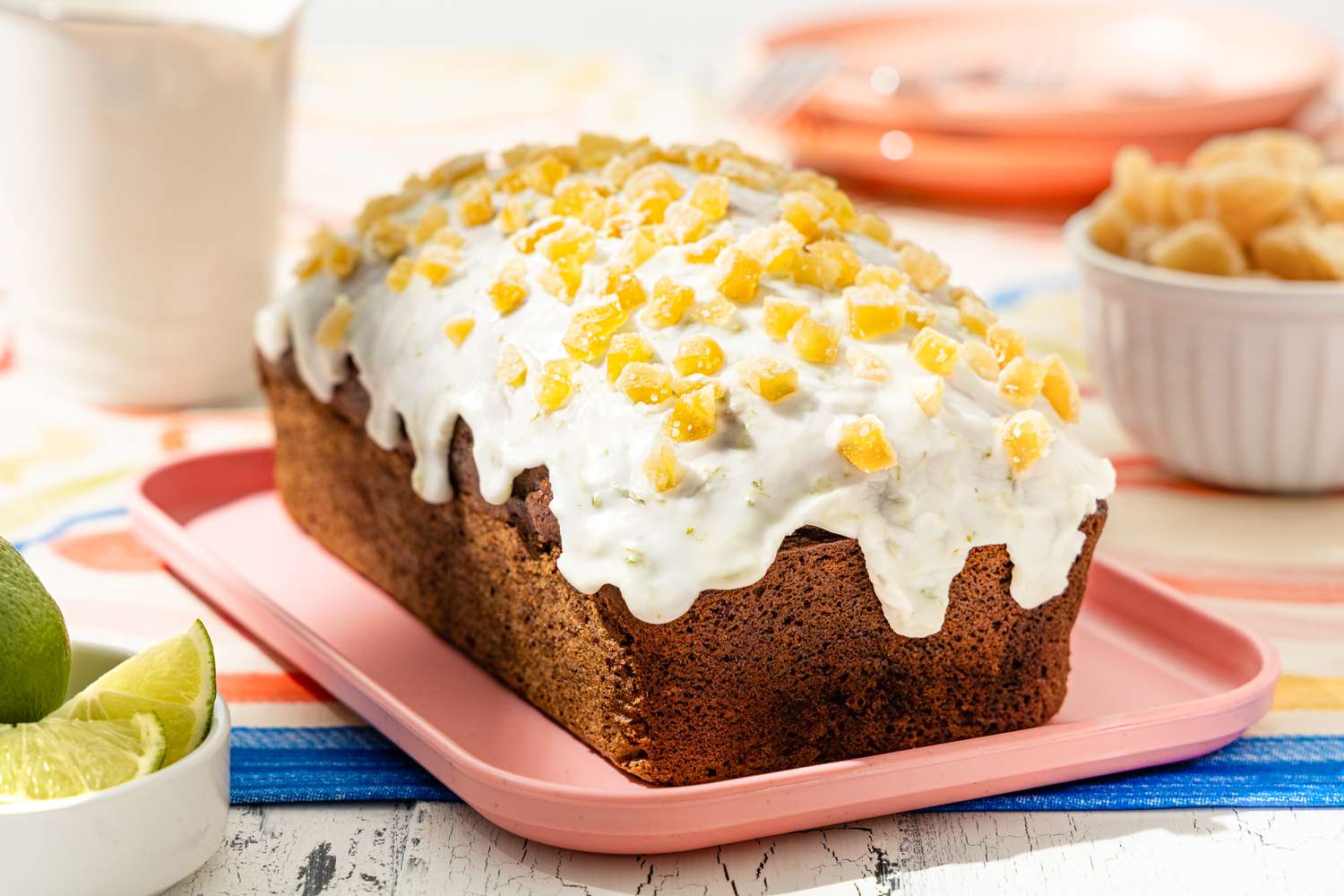 Platter of Dark and Stormy Banana Bread Topped With Glaze and Candied Ginger Pieces, and in the Surroundings, a Bowl of Quartered Lime Slices, Bowl of Candied Ginger, a Pitcher, and a Stack of Pink Plates, All on a Fun Decorative Table Cloth