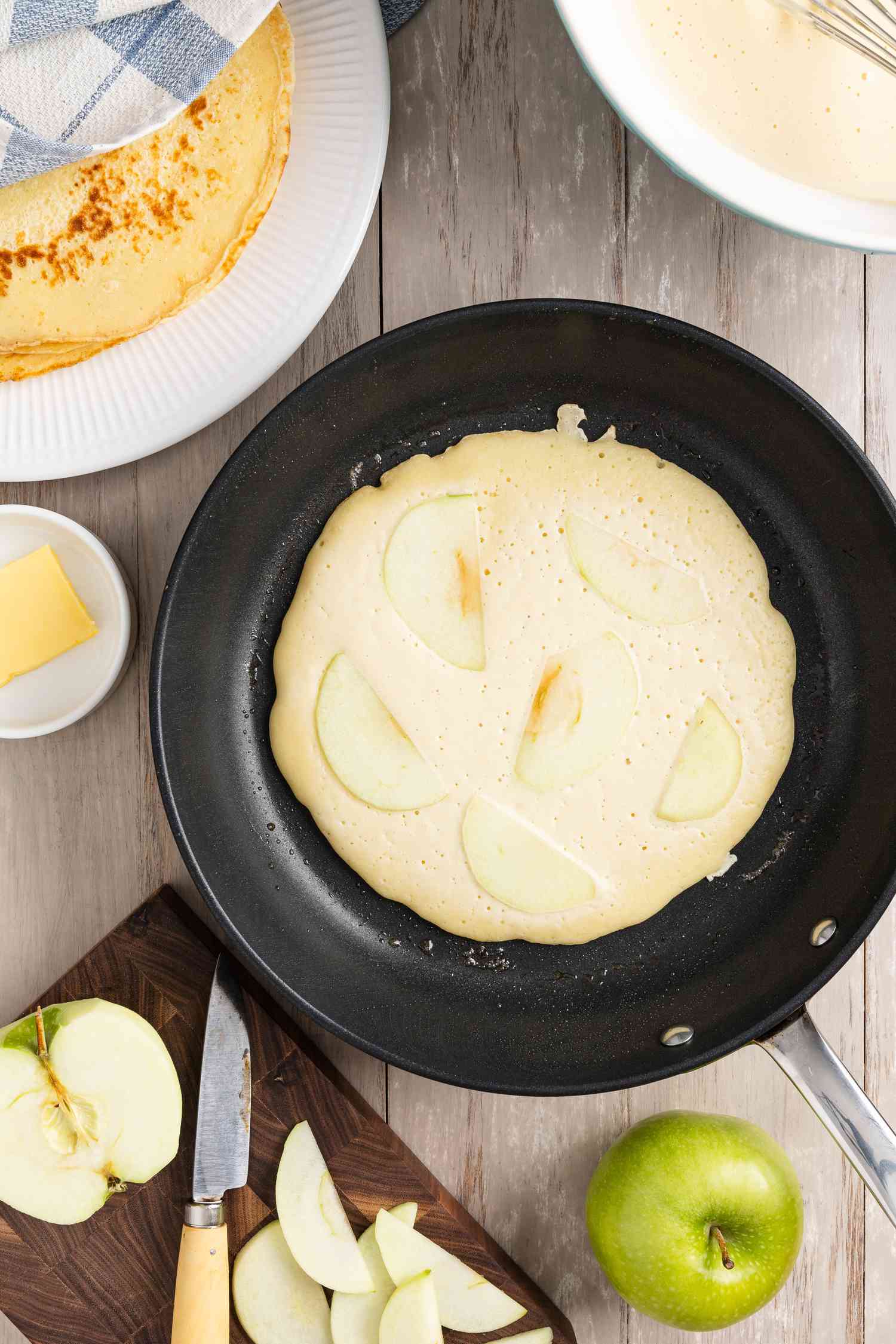 Pannenkoeken (Dutch Pancakes) batter in a skillet topped with apple slices, and in the surroundings, apple slices and half an apple on a cutting board, an apple on the counter, and a plate with more Dutch pancakes