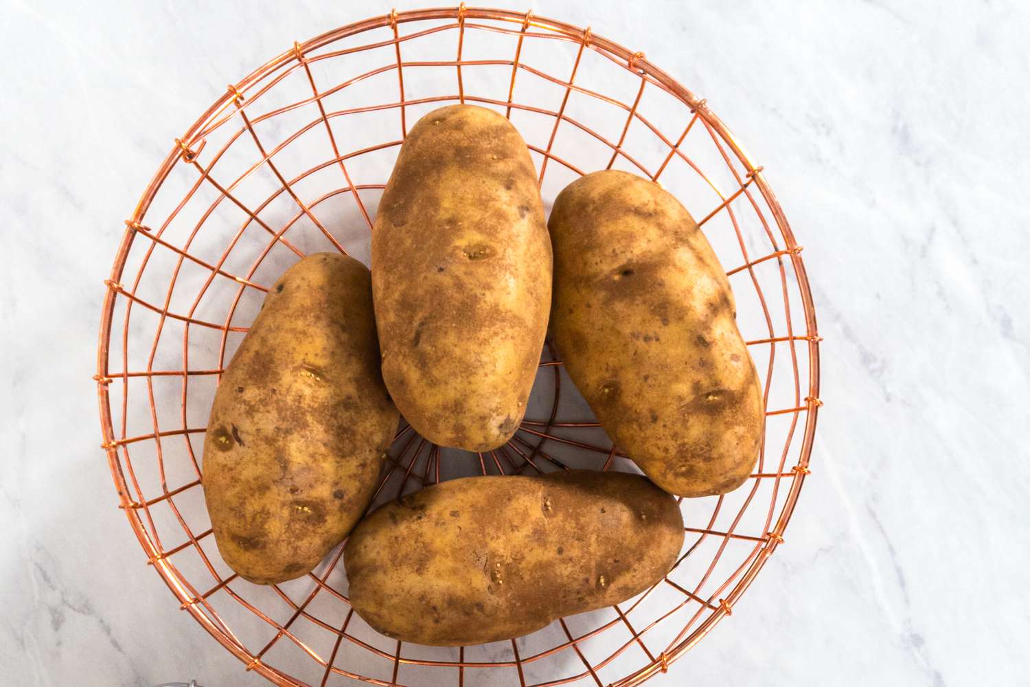 Four raw potatoes in a wire basket on a marble kitchen counter