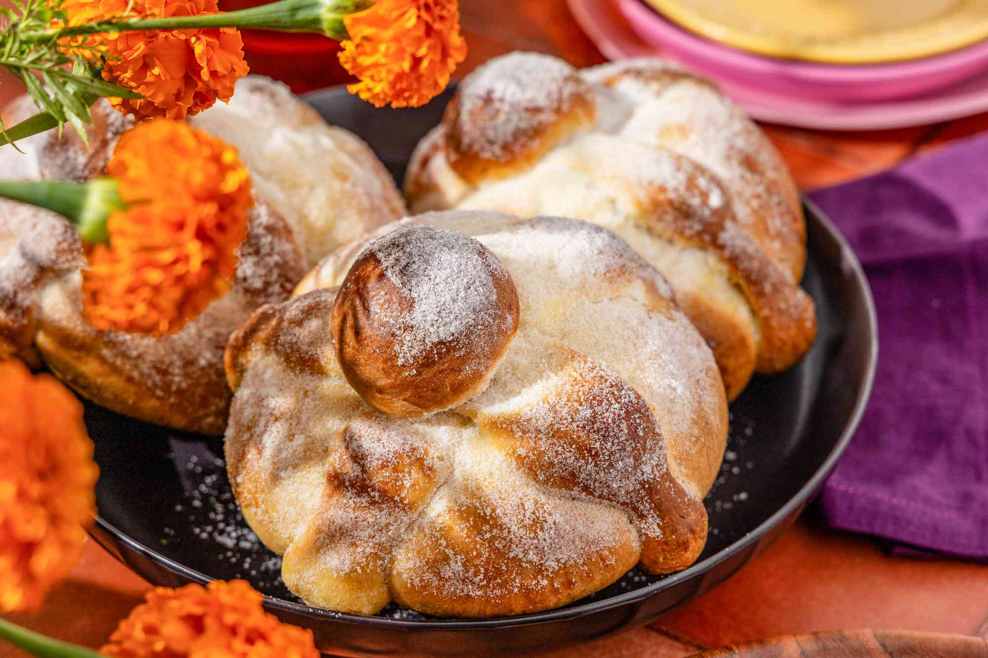 Pan de muerto in a bowl next to some orange carnations, and in the background, a stack of plates, a purple kitchen towel, and glasses of water, all on a brick surface