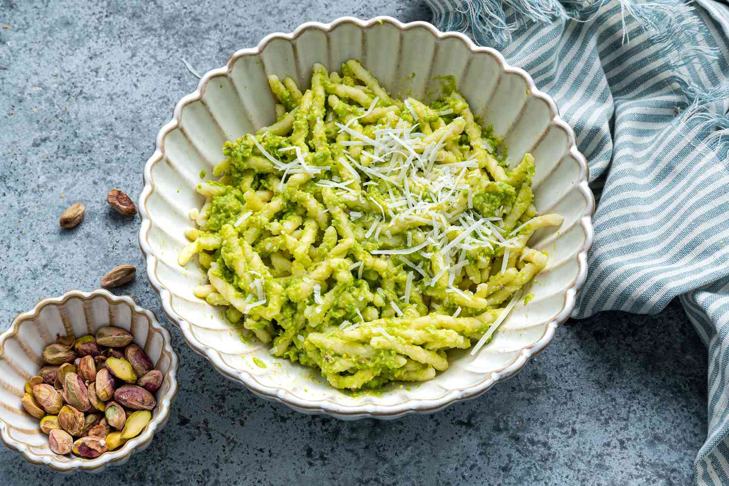 Bowl of Pea Pesto Pasta Topped With Pecorino Romano Cheese Next to a Bowl With Deshelled Pistachios and a Blue and White Stripped Kitchen Towel 