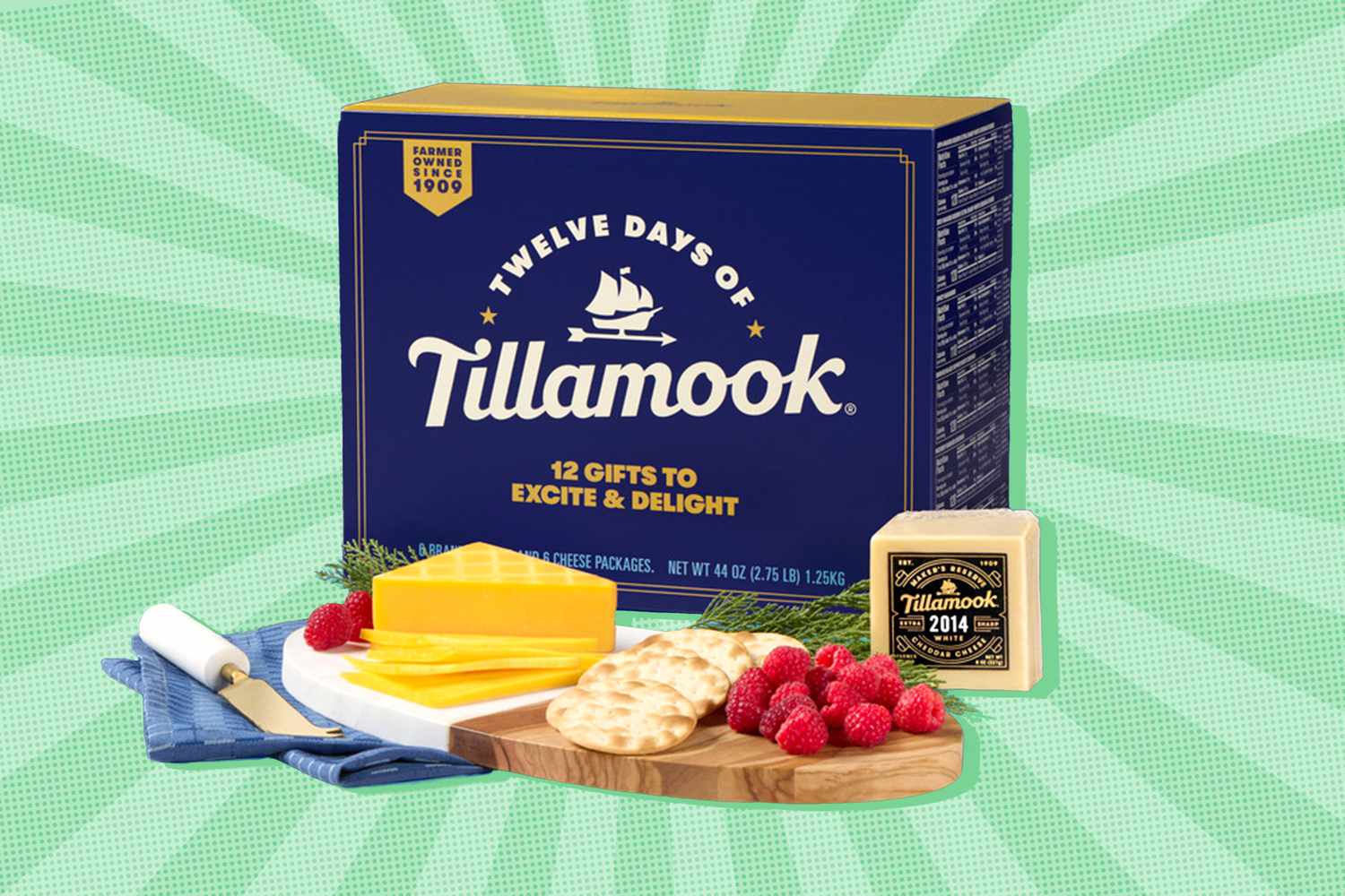 A dark blue box with "Twelve Days of Tillamook" printed on the front. In front of the box is a cheese board with smoked cheddar, water crackers, and raspberries on it.