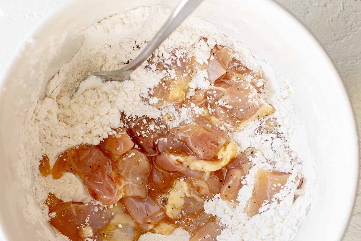 Chopped chicken is being coated in flour to show how to make chinese orange chicken.
