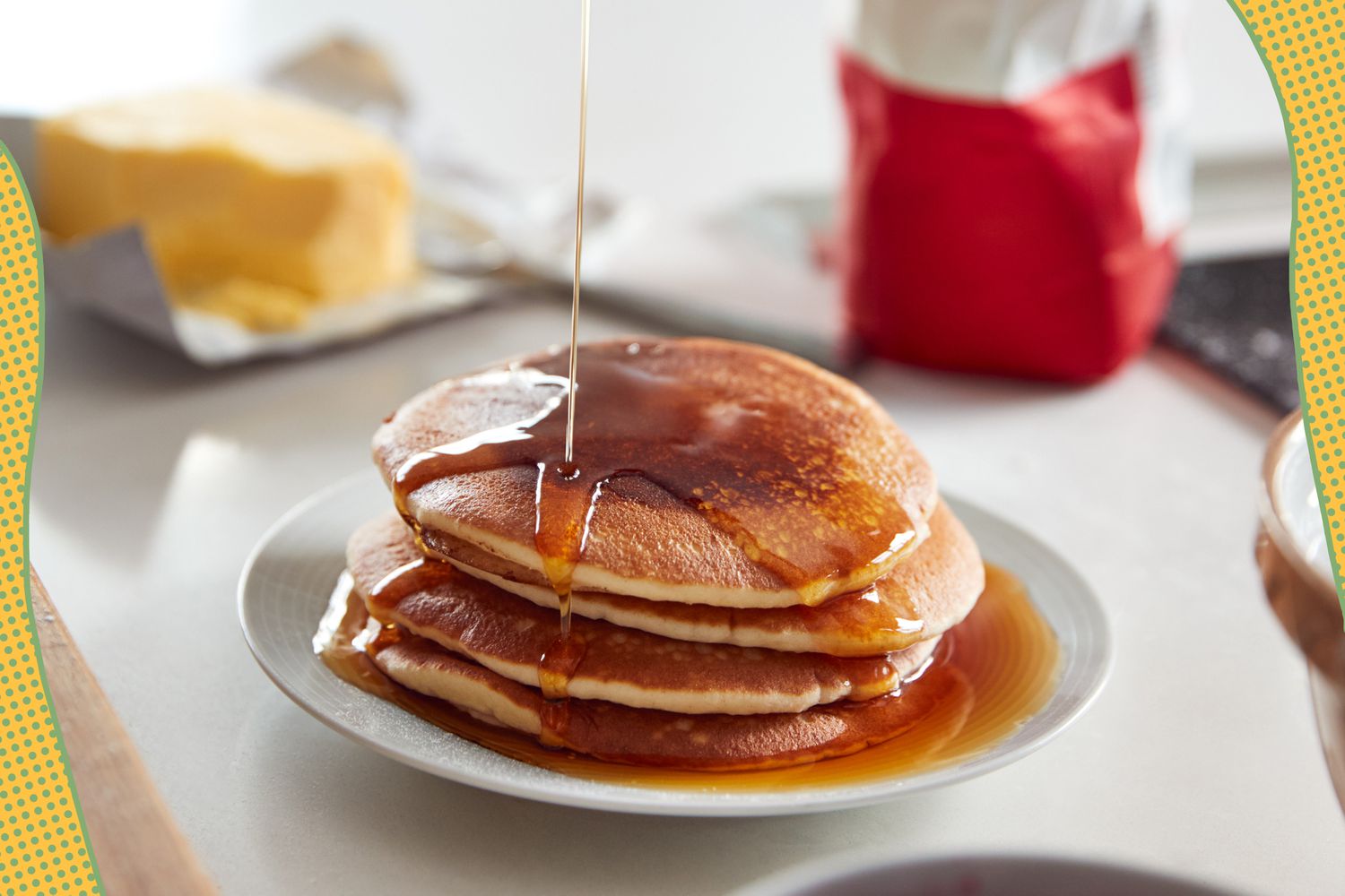 Photo of maple syrup poured onto a stack of pancakes with pancake ingredients in the background, and in the corners of the photo, a yellow wavy color block with blue polka dots