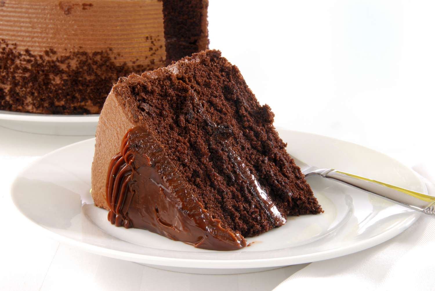 A slice of chocolate layer cake with frosting placed on a white plate with a fork
