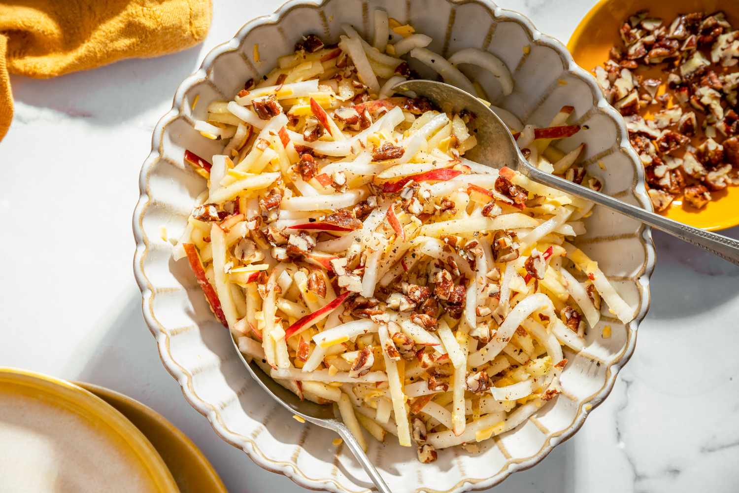 A bowl of kohlrabi and apple salad topped with nuts served with a spoon and garnished with additional nuts nearby