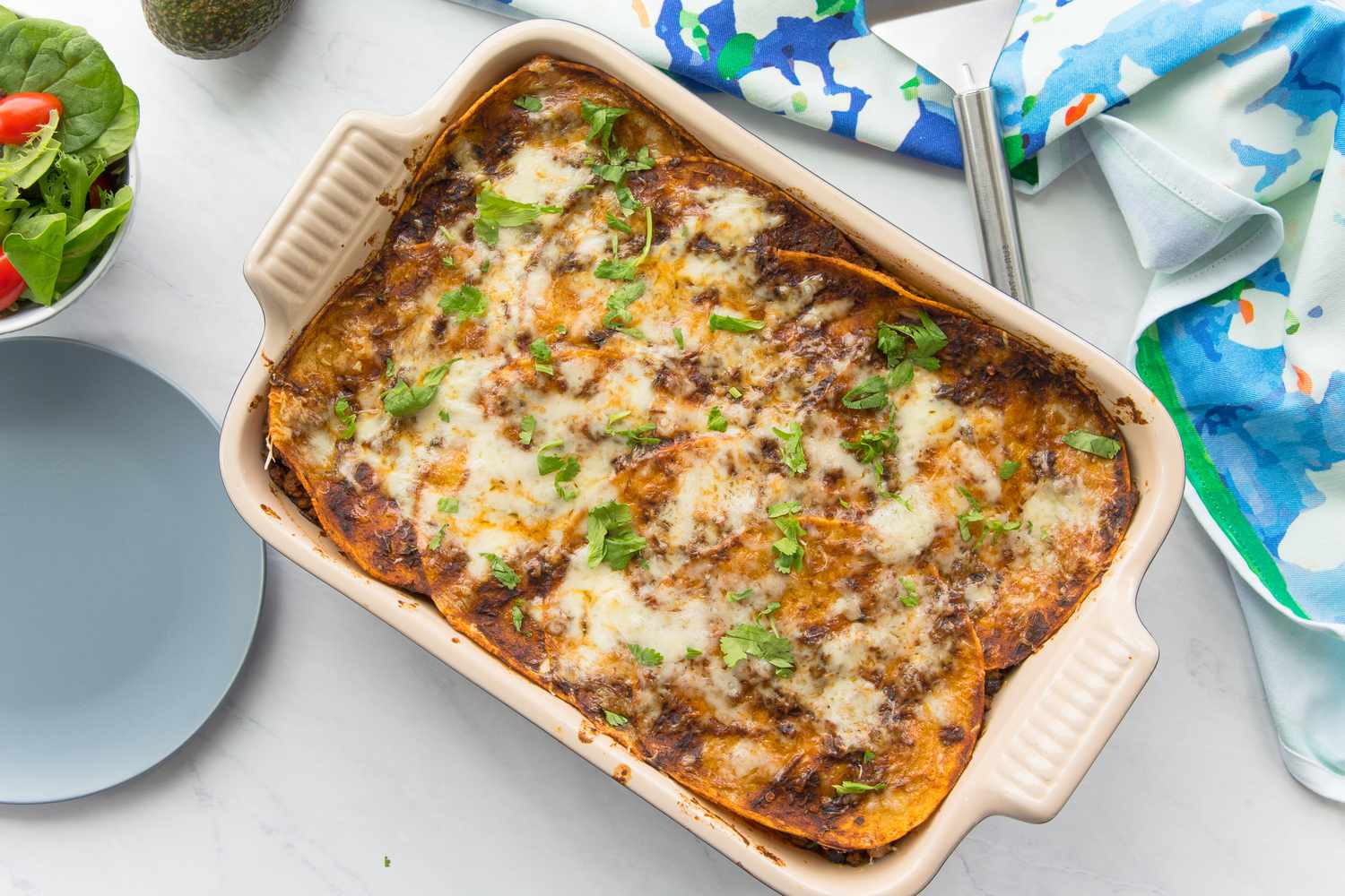 Beef Enchilada Casserole in a Casserole Dish Next to a Table Cloth, a Plate, and a Bowl with Side Salad 