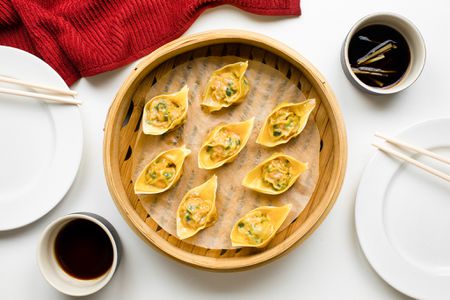 Table Setting: Pork and Chive Dumplings in a Lined Bamboo Steamer, Two Small Bowls of Chinese Vinegar Sauce (One with Sliced Ginger), Two Plates with Chopsticks, and a Kitchen Towel