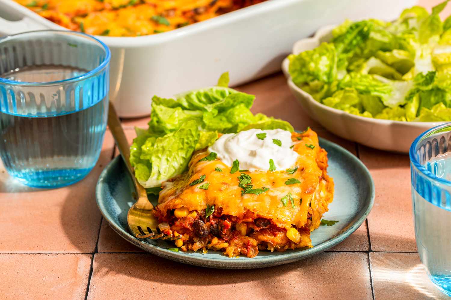 Serving of chicken taco casserole on a plate topped with a dollop of sour cream and served with a side salad, and in the surroundings, glass of water, a casserole dish with more, and a bowl of side salad