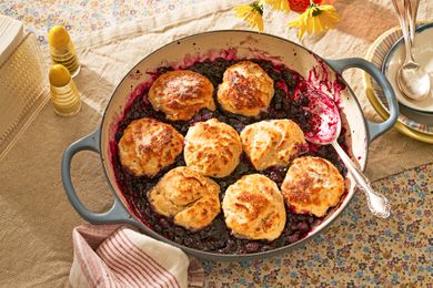 blueberry grunt in a cast iron braiser at cozy looking table with a stack of bowls, salt shakers, a napkin holder with single use napkins, and a vase of flowers