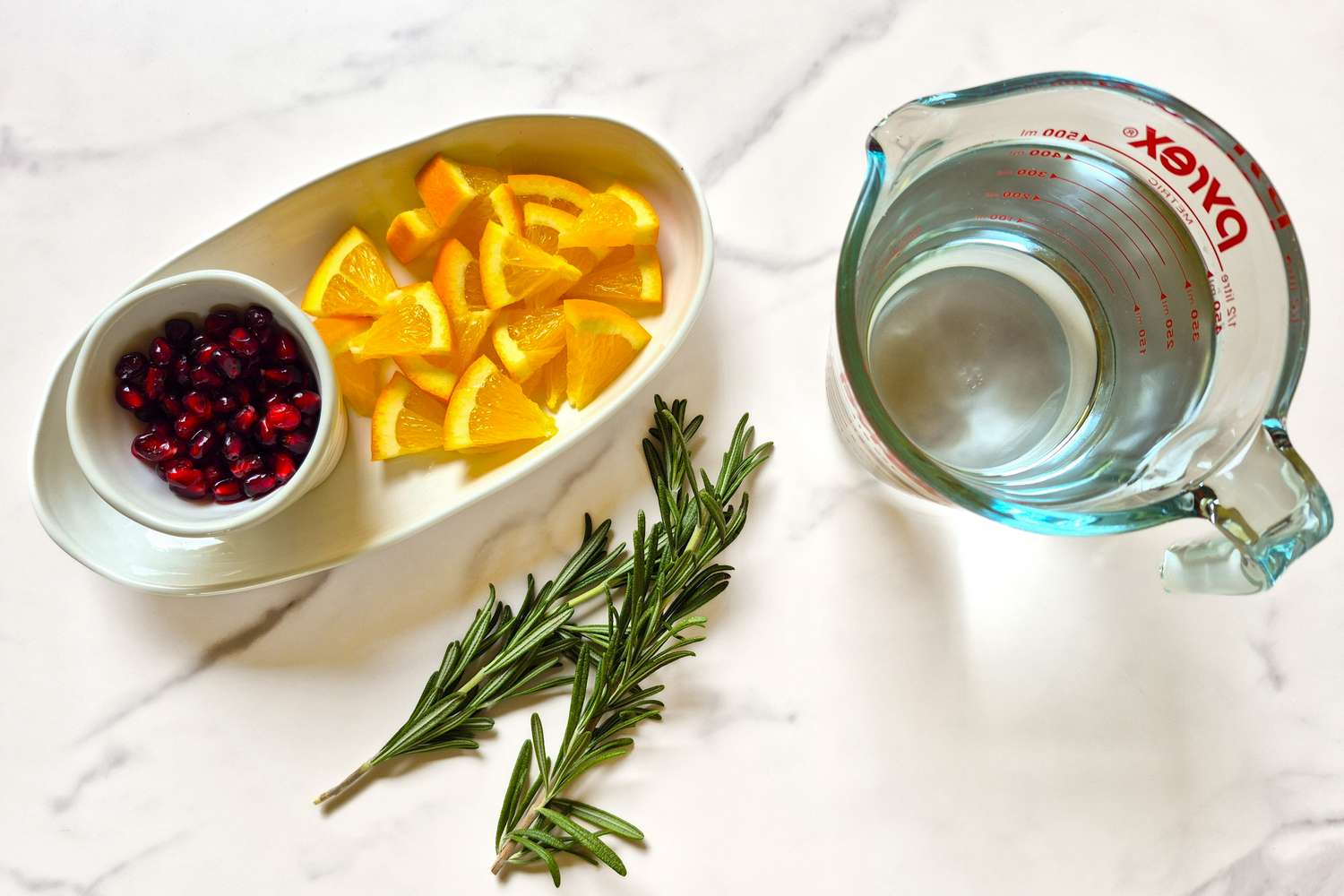 An arrangement of cranberries orange slices rosemary sprigs and a measuring cup of water on a marble surface
