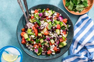 Easy Greek Salad in a Wooden Bowl With a Set of Serving Utensils, and in the Surroundings, a Glass of Iced Lemon Water, a Bowl of Herbs, and a White and Blue Striped Kitchen Towel