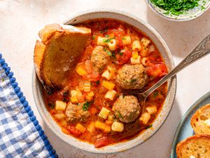 Overhead view of a bowl of Italian meatball soup with a slice of bread and a spoon next to a plate of bread and bowl of parsley
