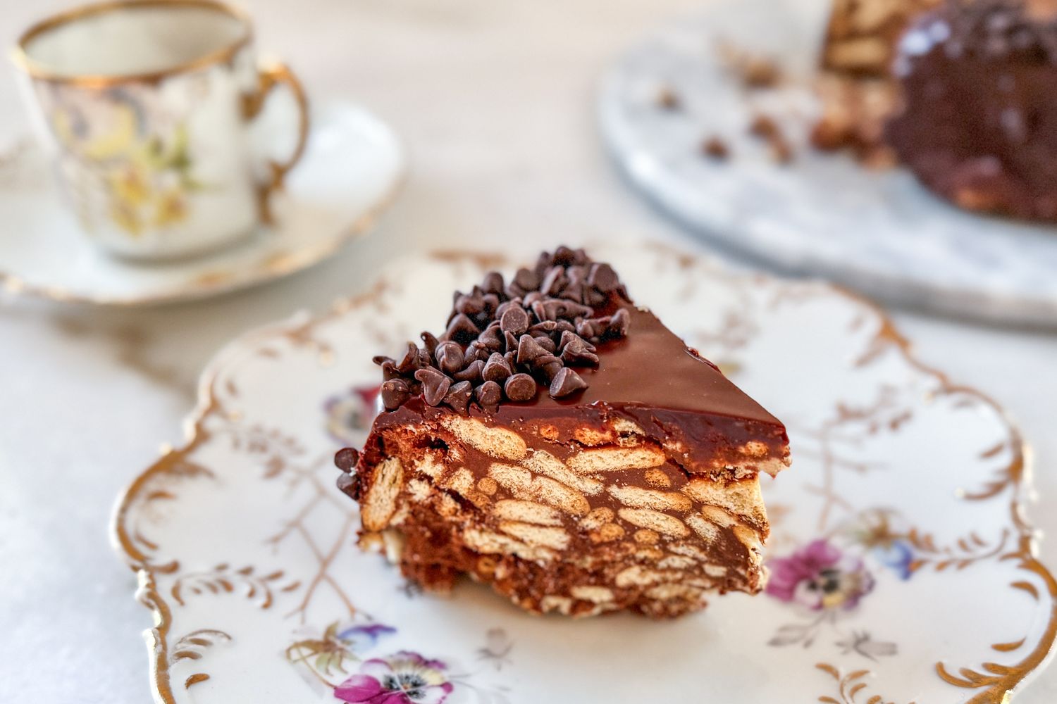 A slice of chocolate nobake cake with biscuit chunks displayed on a decorative plate with a teacup and more cake in the background