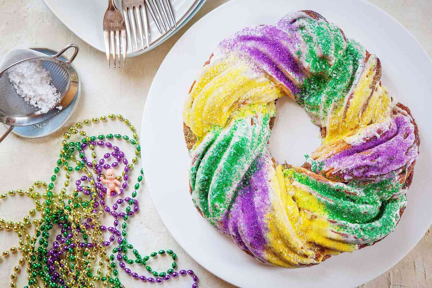 Top view of the best king cake on a white platter. Mardi Gras beads, a sifter of powdered sugar and a stack of plates and forks are to the left.