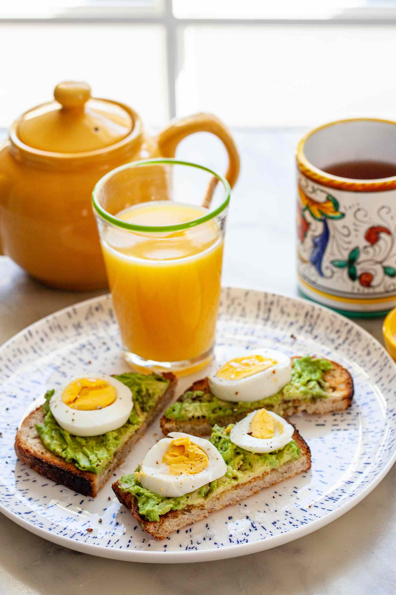 Hard Boiled Eggs on Avocado Toast Next to a Glass of Orange Juice, Teapot, and Mug