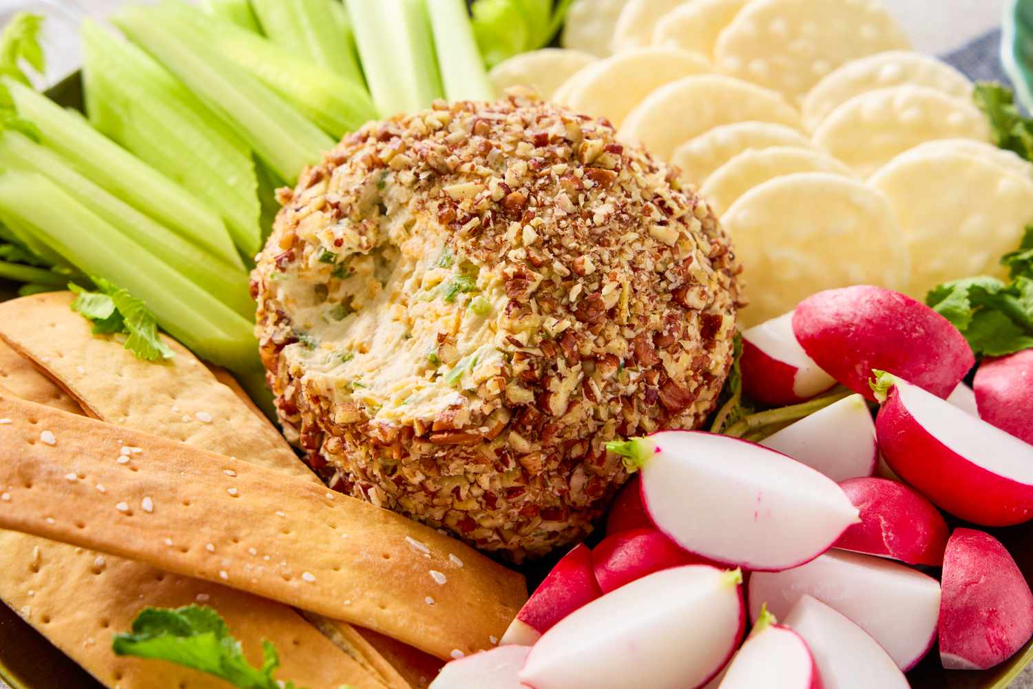 A cheese ball coated in nuts surrounded by crackers radishes celery and other snacks
