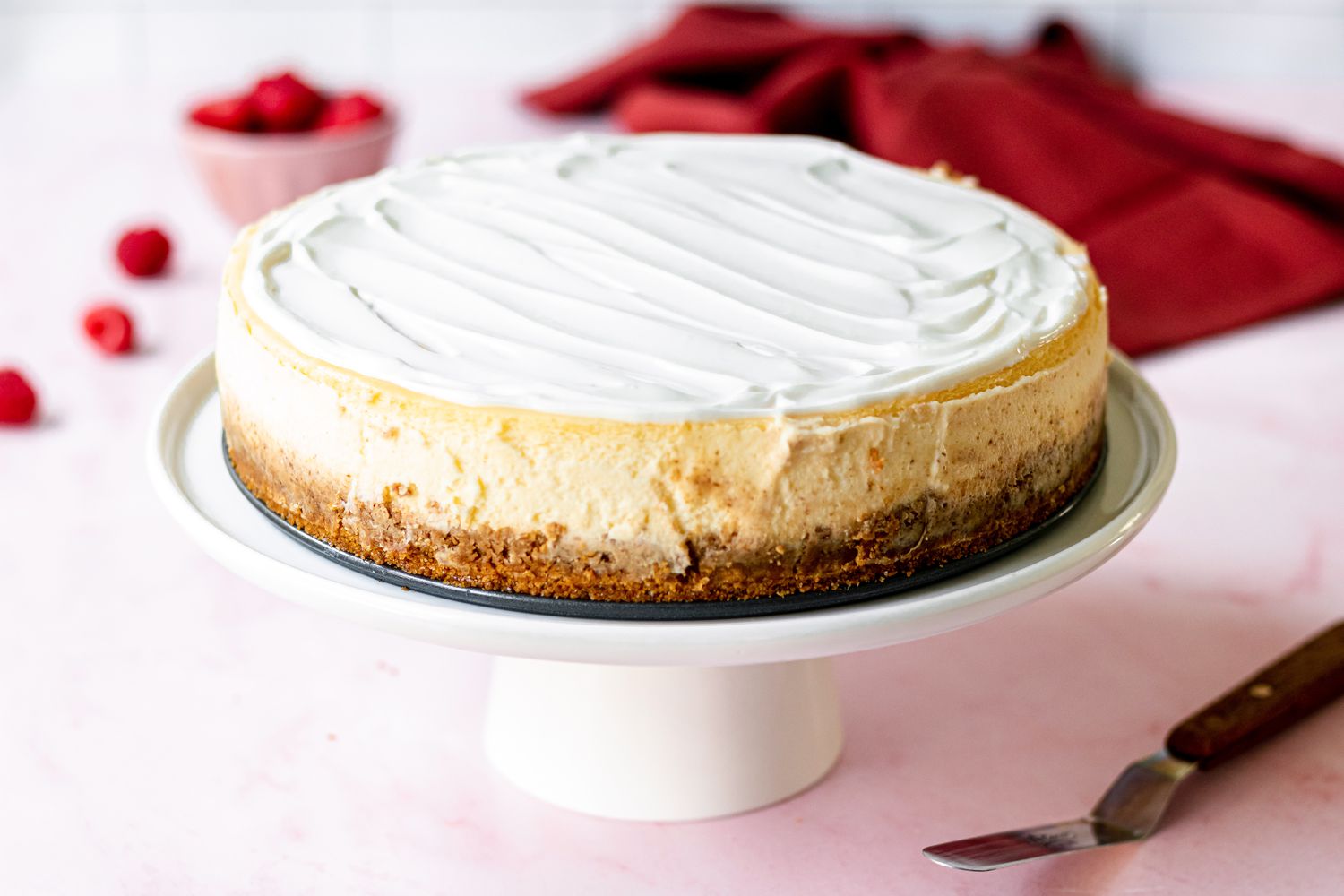 A whole cheesecake set on a cake stand with raspberries behind it.