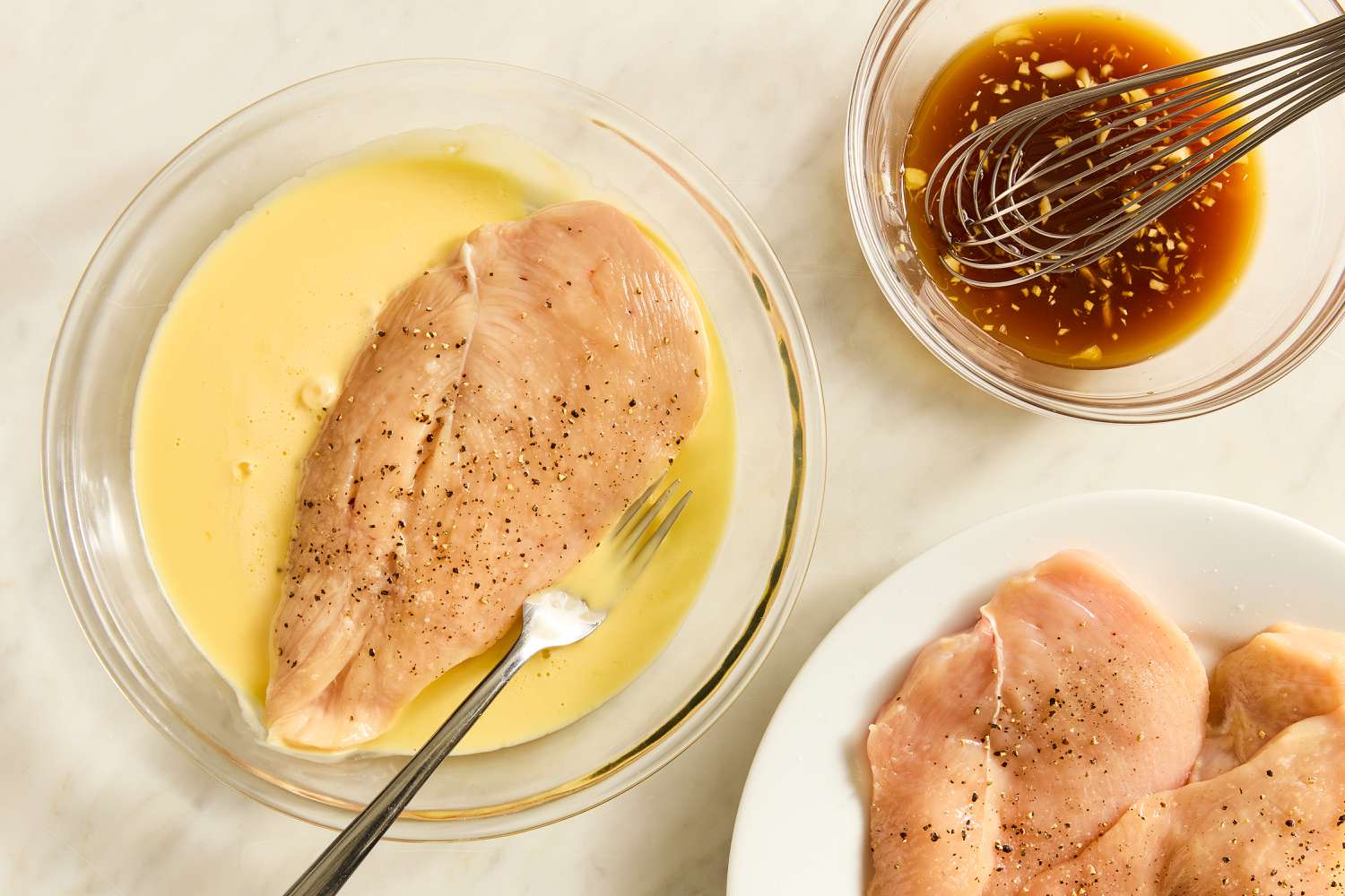Overhead view of a clear glass bowl of with a raw chicken breast in egg mixture next to a bowl of soy sauce mixture and a plate of raw chicken for Honey Soy Chicken recipe