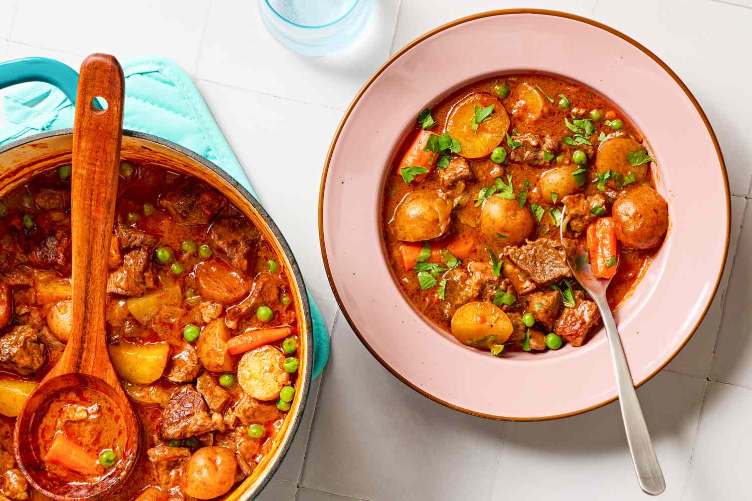 Overhead view of a teal colored dutch oven of hands-off beef stew and a wooden spoon next to a pink bowl of stew with spoon and all on a tile countertop
