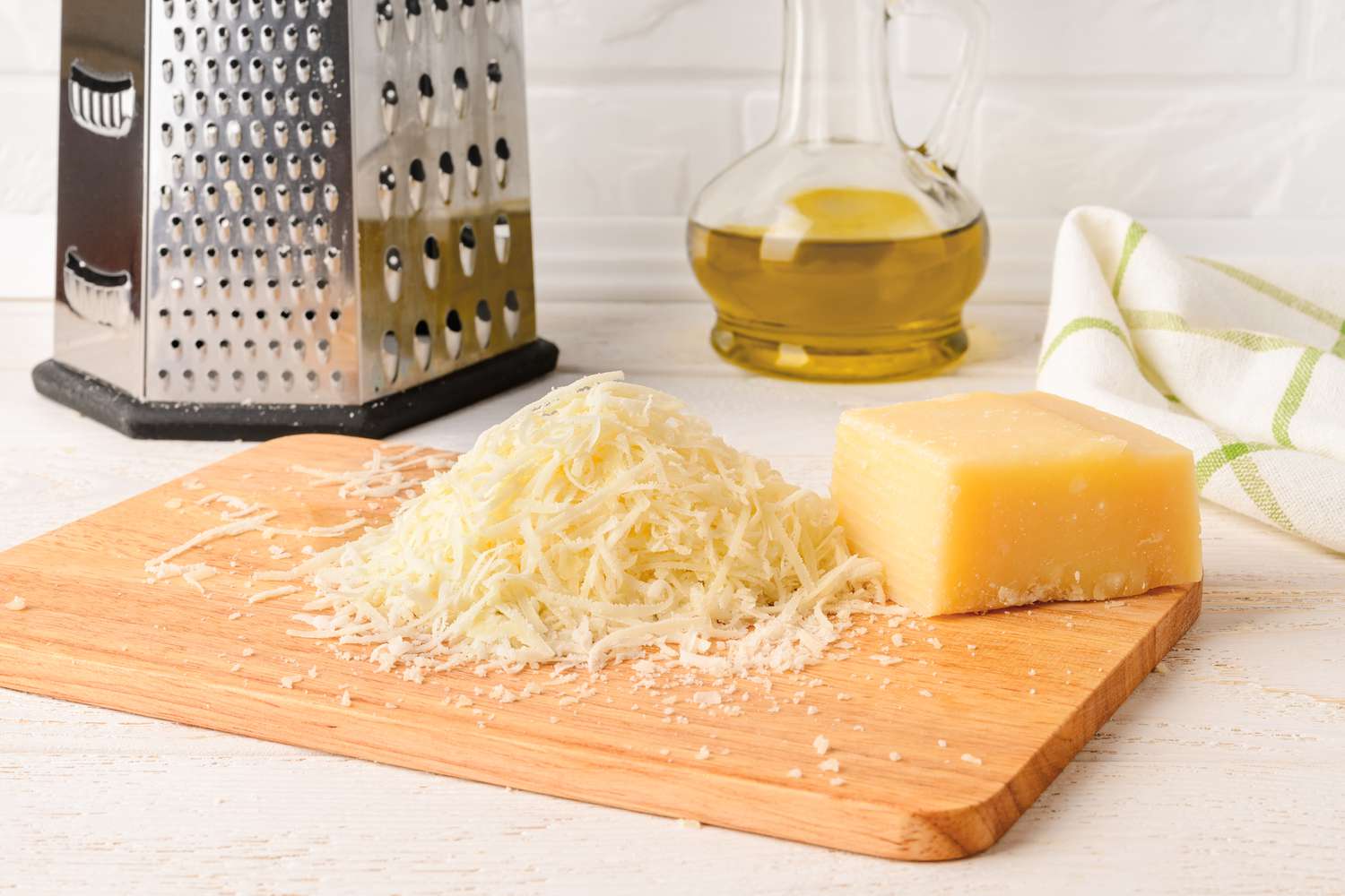 shredded parmesan and a block of parmesan on a cutting board and in the background, a box grater and a carafe with olive oil