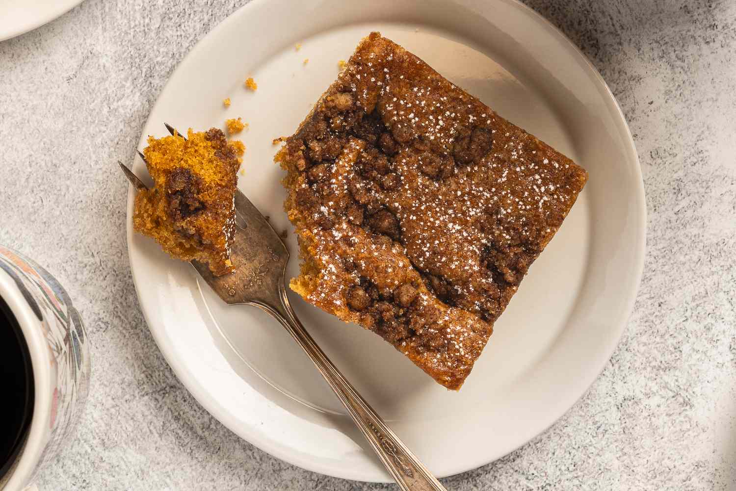 A slice of coffee cake with powdered sugar and crumble topping on a plate with a fork holding a bite