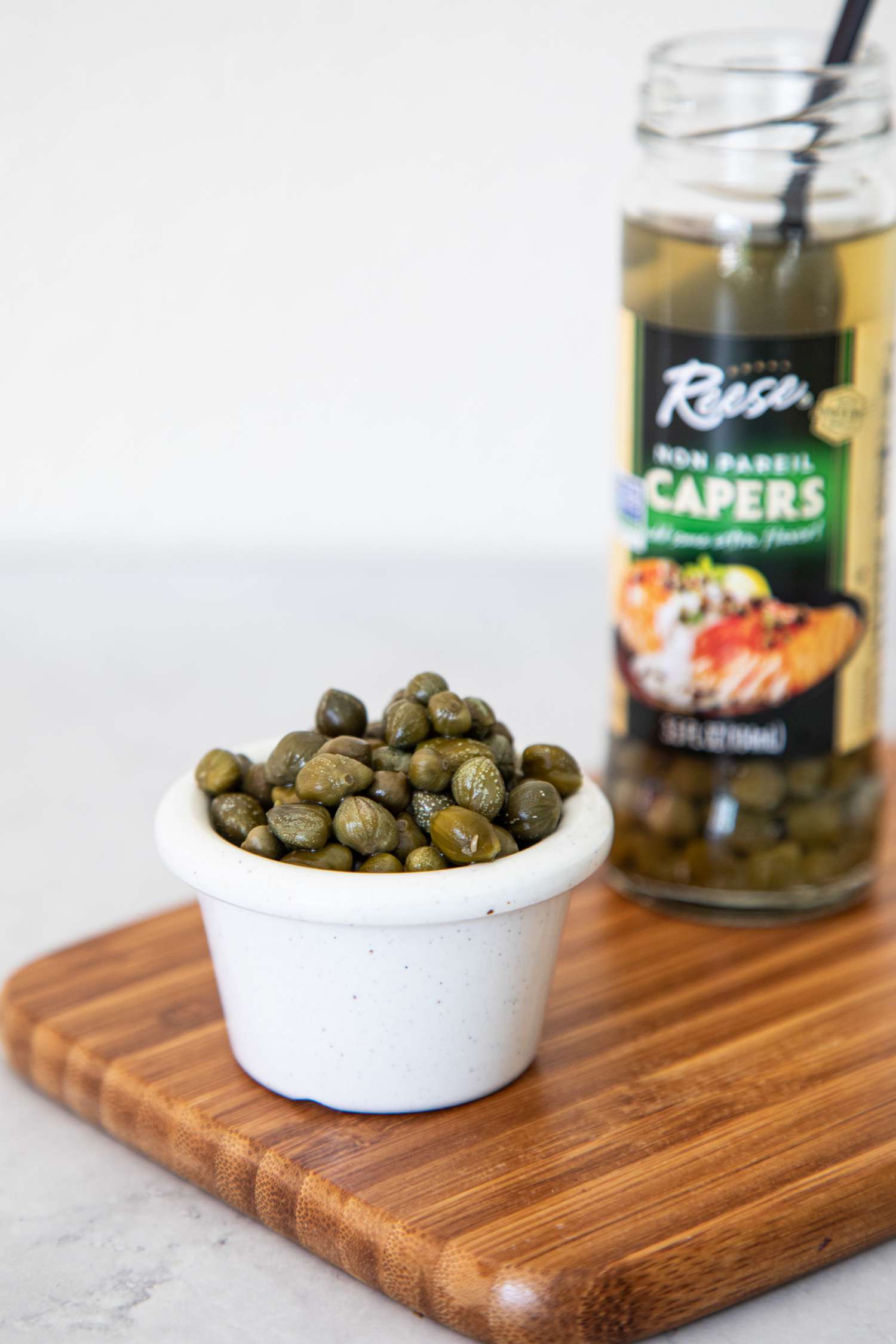 Small Bowl of Capers with the Jar Behind It, and Both Are on a Wooden Board