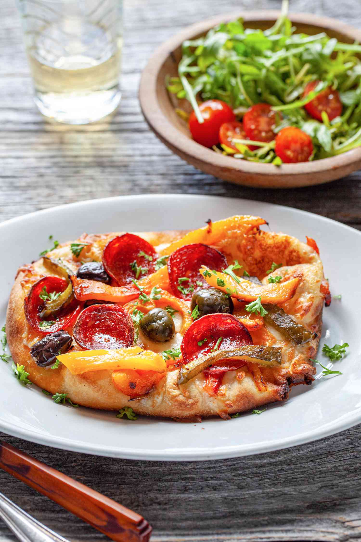 Personal Pizza Topped with Pepperoni, Olives, and Grilled Peppers Next to a Bowl of Salad