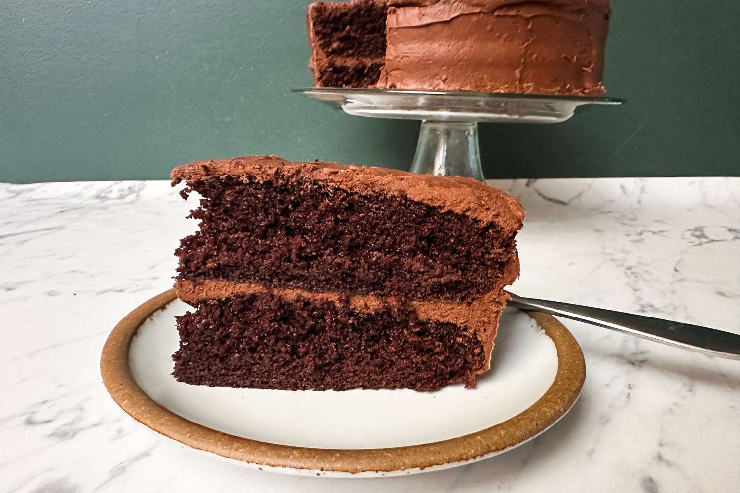 A slice of chocolate layer cake on a plate with the rest of the cake on a stand in the background