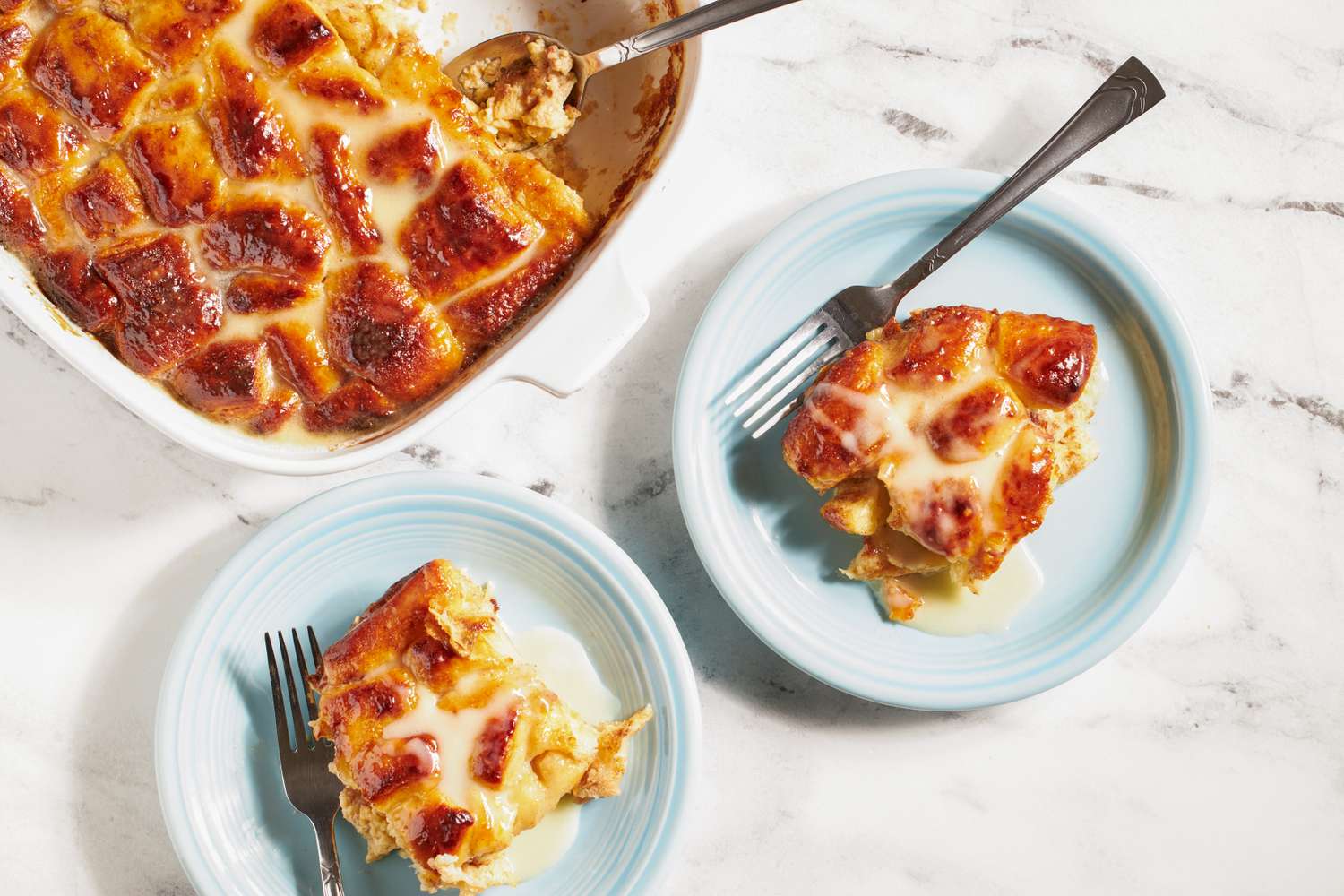 Baking dish with cinnamon roll bread pudding and two plates serving portions of it set on a marble surface