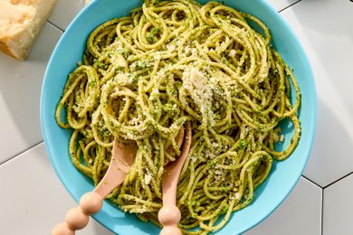 A bowl of pasta coated with pesto garnished with grated cheese with wooden utensils placed on top