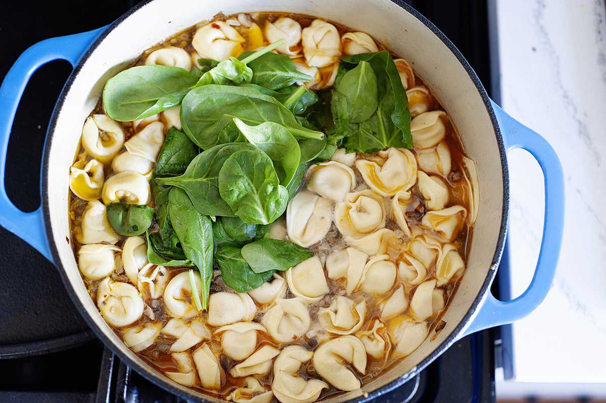 Dutch oven with the best tortellini soup simmering on the stove. Spinach is on top of the tortellini before being mixed into the soup.