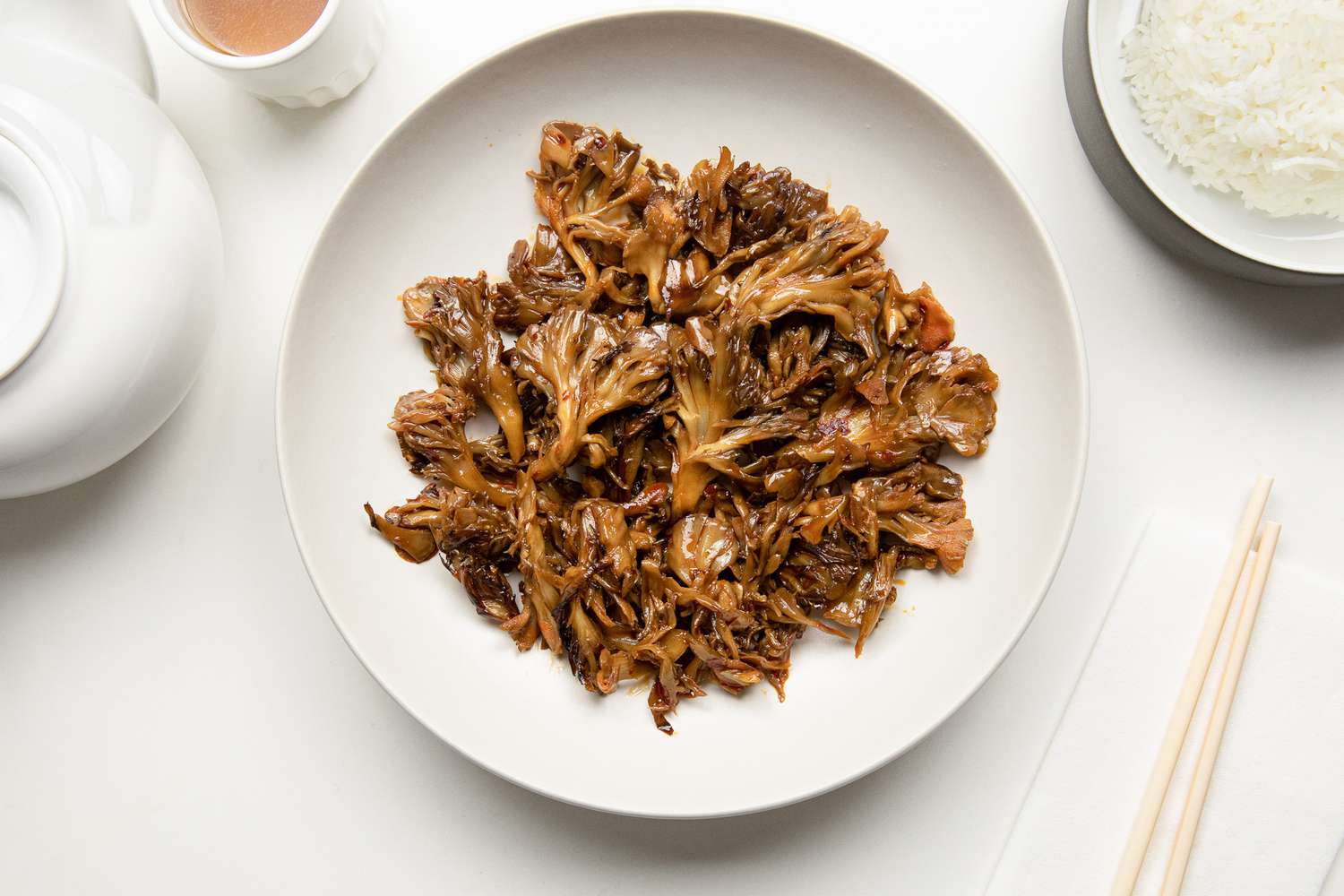 Stir-Fried Maitake Mushrooms with Garlic and Chile Oil in a Plate Next to a Pair of Chopsticks, a Bowl of Rice, a Cup of Tea, and a Tea Kettle