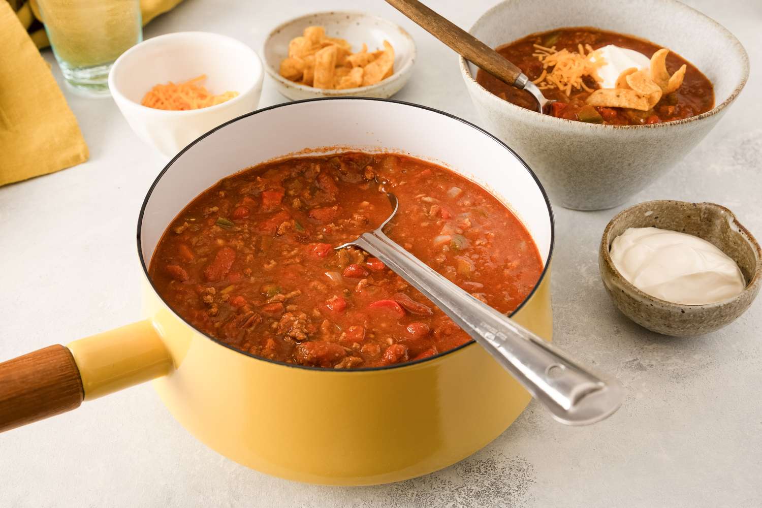 A pot of Grandma Patty’s Chili with a serving spoon with bowls of chili and sides in the background