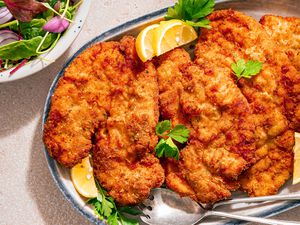 Chicken Romano on a platter, garnished with flat-leafed parsley and lemon wedges, bowl with mixed salad greens on the side
