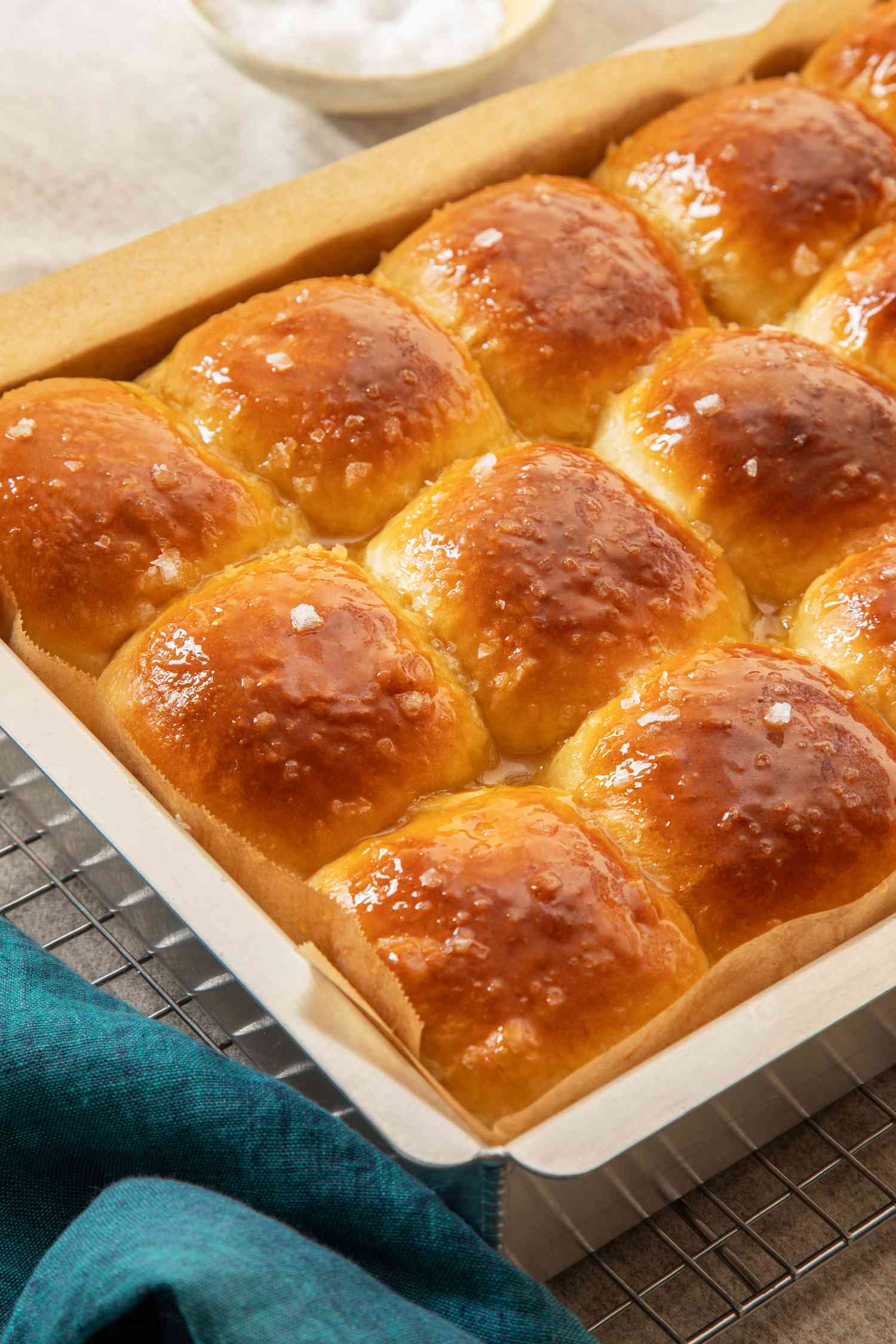 Salted honey rolls in a parchment paper-lined baking sheet on a cooling rack next to a blue kitchen towel
