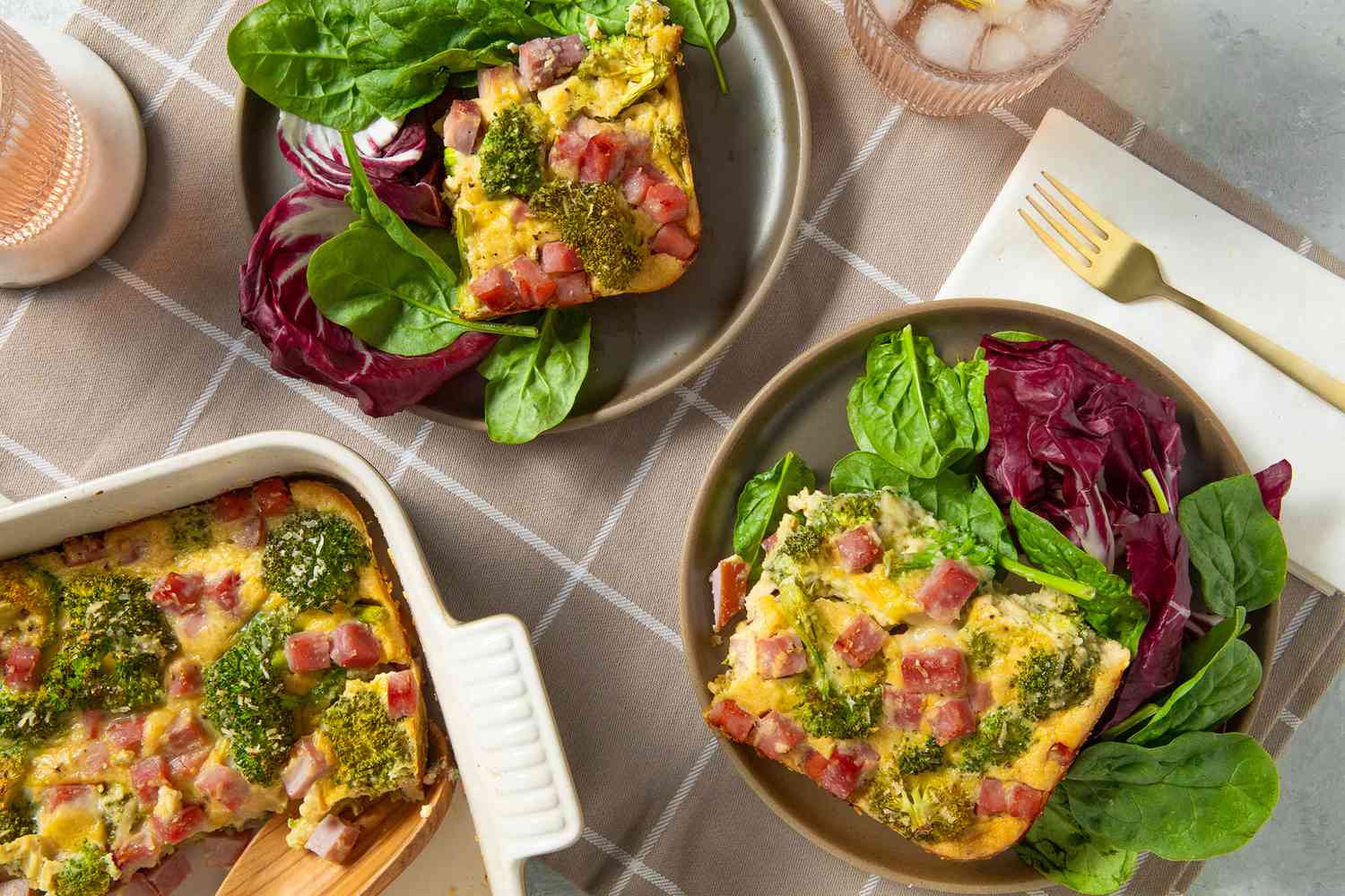 Overhead view of two plates with slices of broccoli cheese pie and greens in a table setting with forks, napkin, drink glasses and a corner of the casserole dish visible