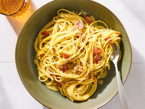 A plate of pasta carbonara with grated cheese and bits of meat with a fork and a glass of beverage nearby