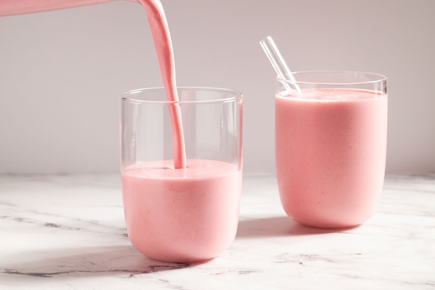 Two glasses of pink smoothie on a marble countertop with smoothie being poured into one glass a straw in the other glass
