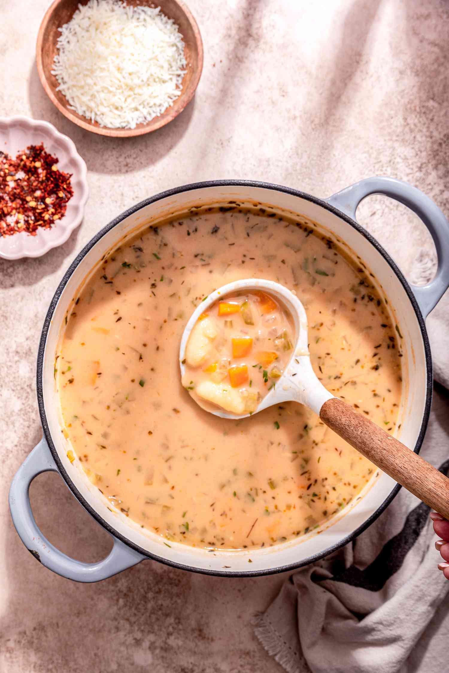 Ladle full of creamy vegetable dumpling soup lifted from a Dutch oven with more, and on the counter surroundings the pot, a bowl of crushed peppers, a bowl of shredded parmesan, and a grey and black kitchent towel