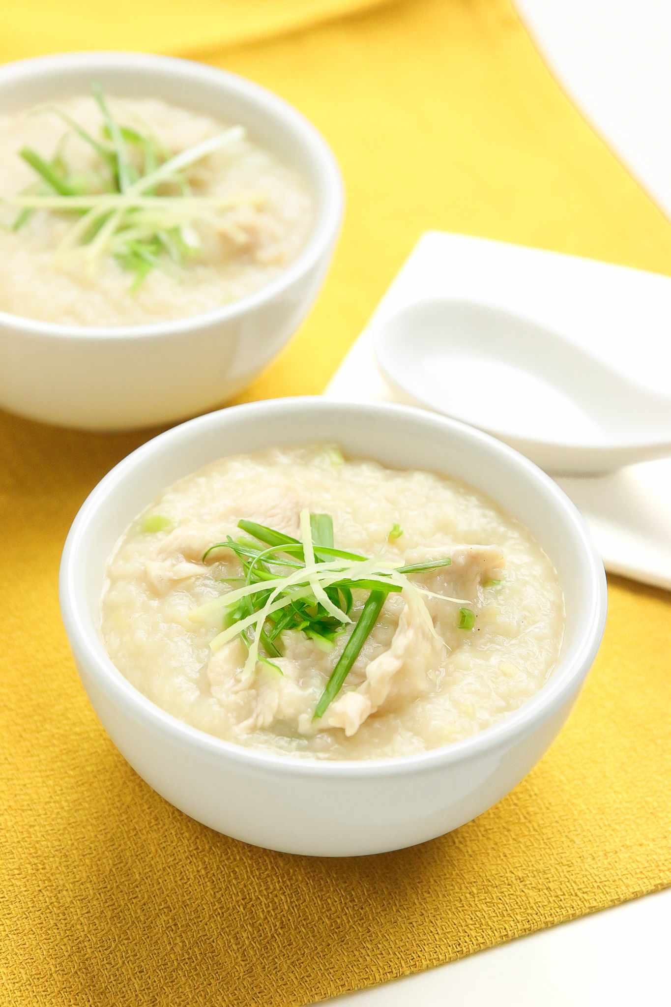 Two Bowls of Chicken Congee Topped with Sliced Ginger and Sliced Green Onion, and Next to the Bowls, Two Soup Spoons on Table Napkins, All on a Mustard Colored Kitchen Towel