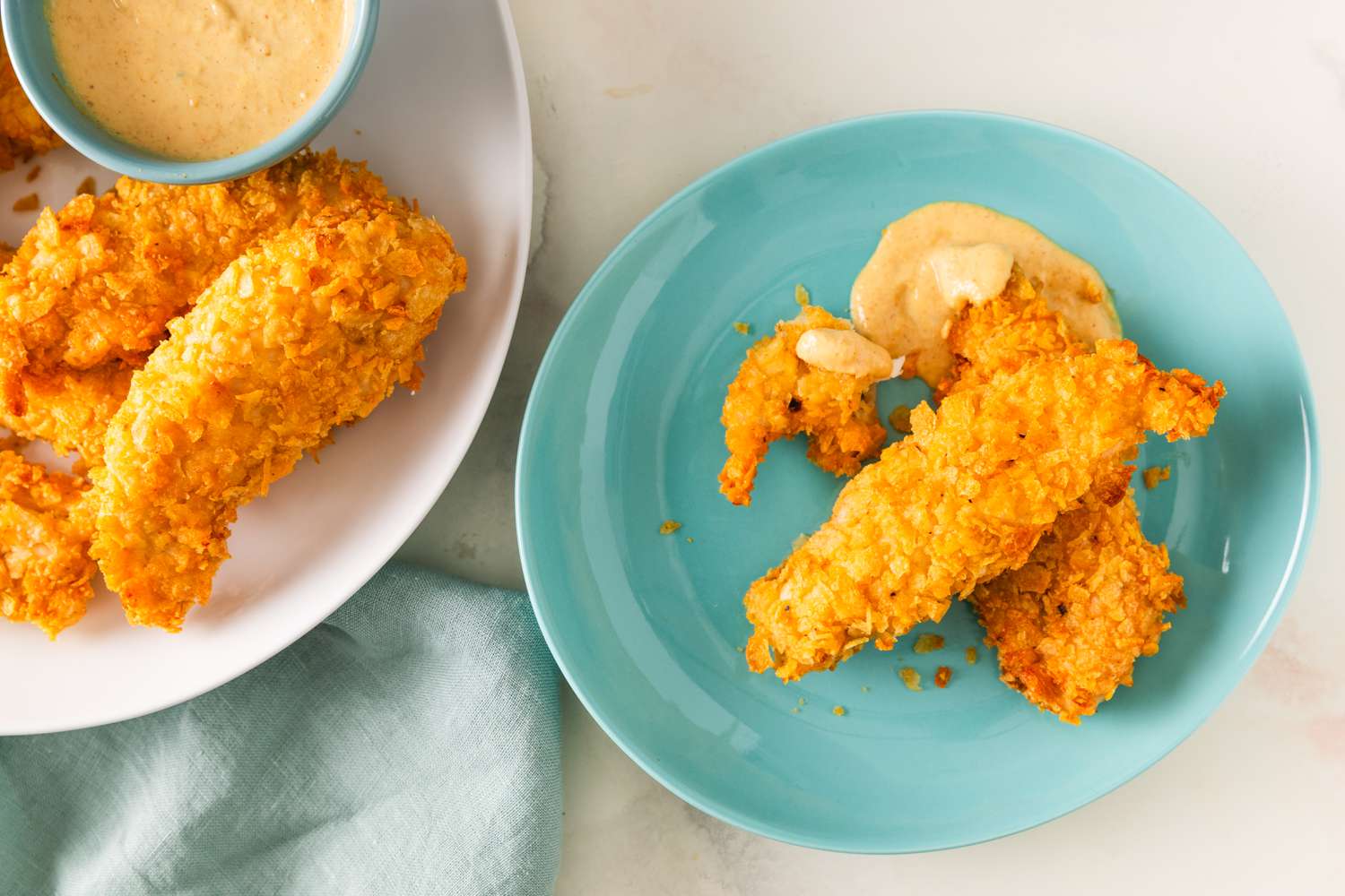 Overhead view of a small blue plate of air fryer potato chip chicken tenders and sauce next to a white plate of tenders and a small bowl of dipping sauce
