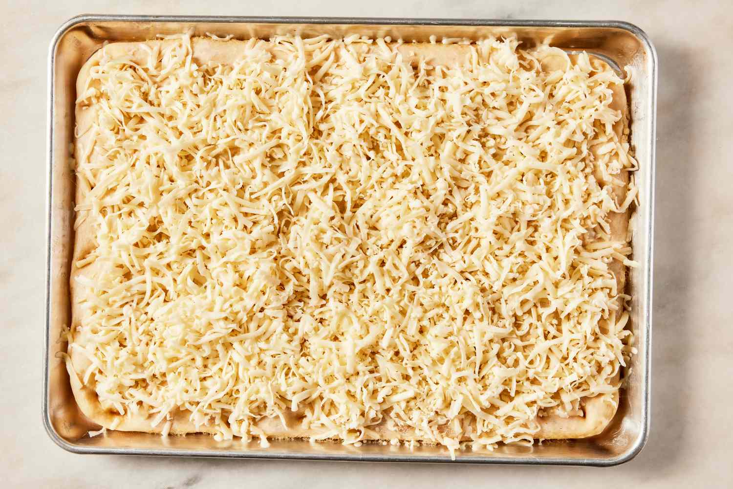 shredded mozzarella added to parbaked grandma pizza dough in a sheet pan