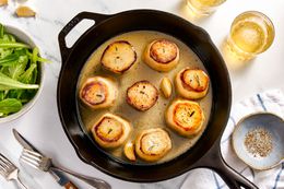 Fondant potatoes in a cast iron skillet at a table setting with a bowl of side salad, utensils on the counter, a bowl of pepper on a kitchen towel, and two glasses