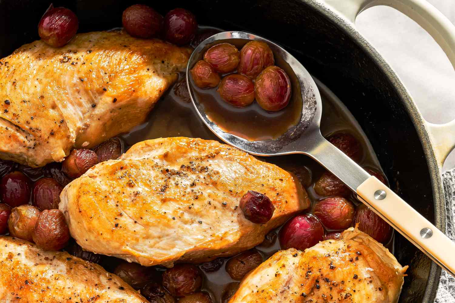 Cooked chicken breasts in a pan with grapes and balsamic sauce a serving spoon holding grapes and sauce