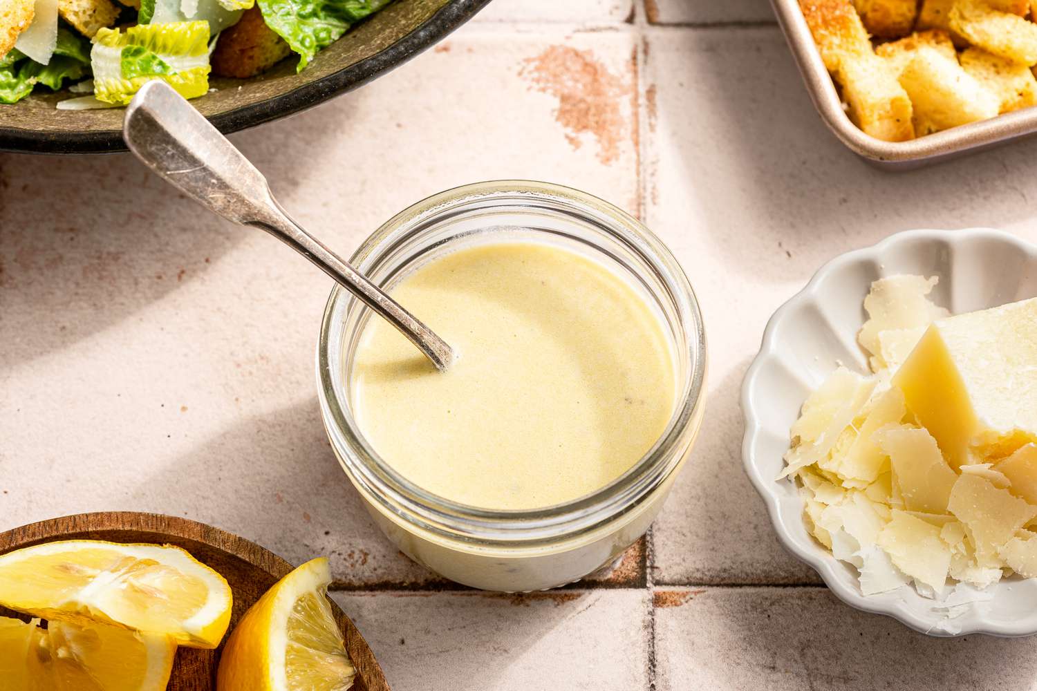 Caesar salad dressing in a jar with a spoon and Parmesan, lemon wedges, and salad greens on the side