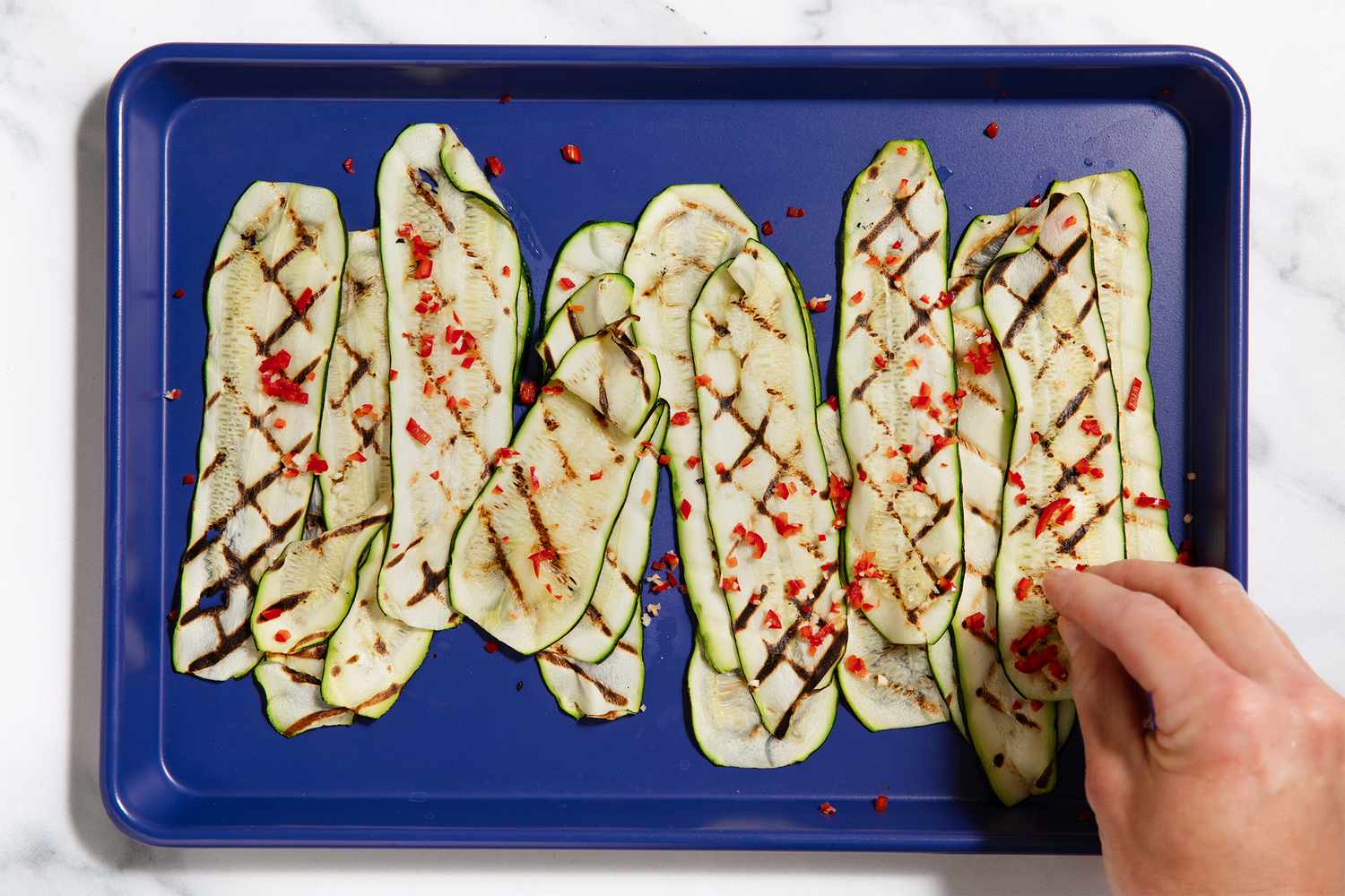 Thin slices of grilled zucchini arranged on a blue tray, garnished with seasoning and a hand placing an additional piece